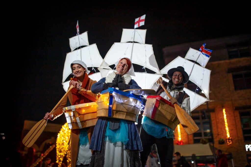 Three Admirable Admiral performers at night wearing boat-shaped props around their waists, decorated with small lights. Each performer holds a wooden oar and stands in front of large white sail structures with flags on top. Festive lights and a building are visible in the background.