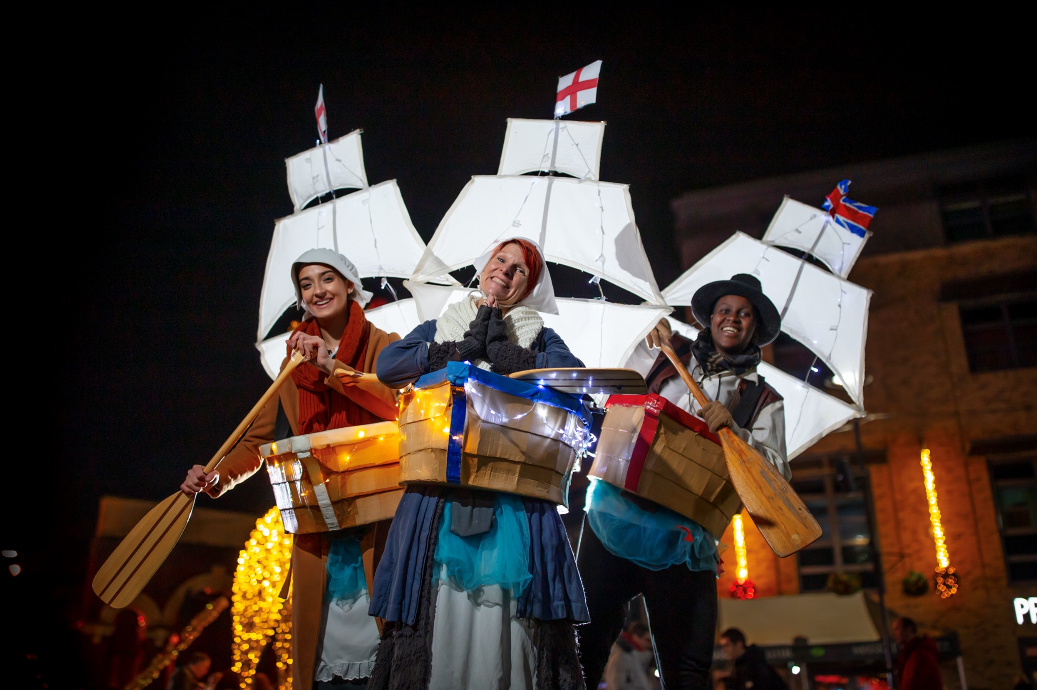 Three Admirable Admiral performers at night wearing boat-shaped props around their waists, decorated with small lights. Each performer holds a wooden oar and stands in front of large white sail structures with flags on top. Festive lights and a building are visible in the background.