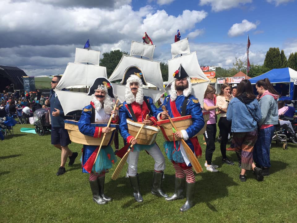 Three Admirable Admirals performers standing on grass at a daytime outdoor event, wearing costumes with large boat props around their waists and holding oars. Behind them are tall sail structures with small flags. A crowd of people, tents, and a stage area are visible in the background under a partly cloudy sky.