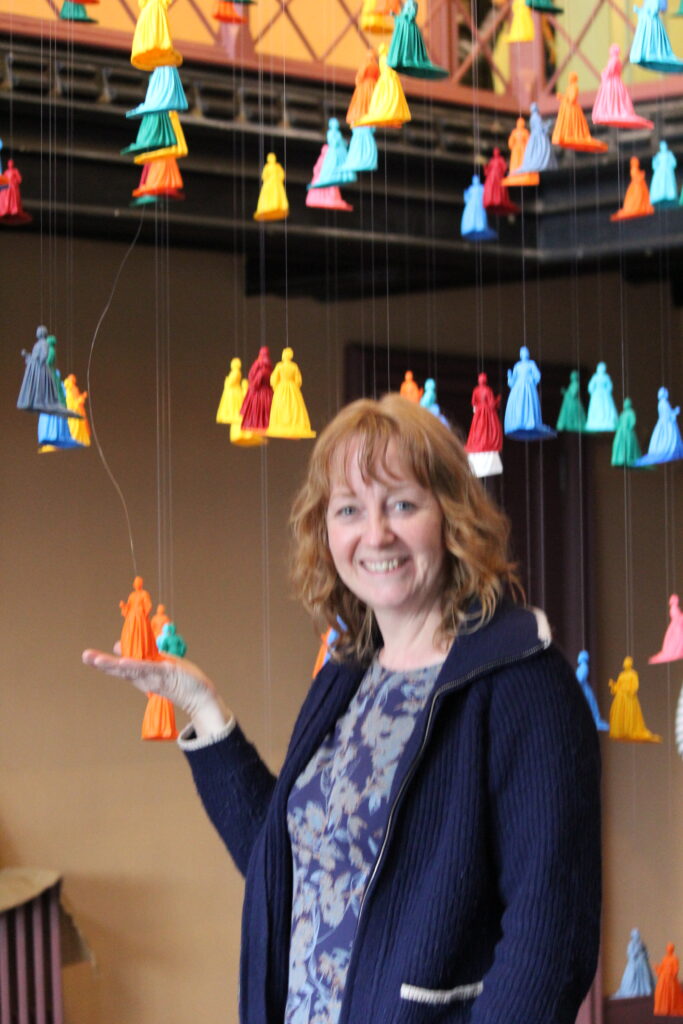 Artist Rachel Carter in front of her colourful suspended 3D figures in Pilgrim Woman Assembly installation at Shodfriars Hall Boston
