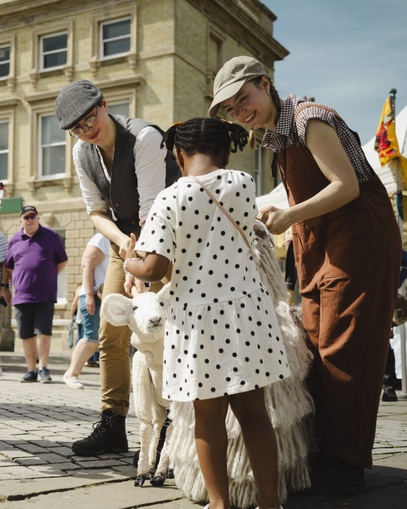 Child interacting with a Lincolnshire Longwool sheep and lamb puppets during a family-friendly outdoor arts event in a town square