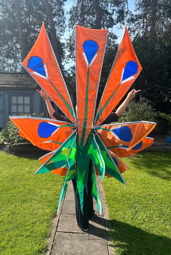 Artist Ruth Pigott wearing bright orange skullcap flower costume outdoors, showing Hanse 2026 parade design inspired by local heritage