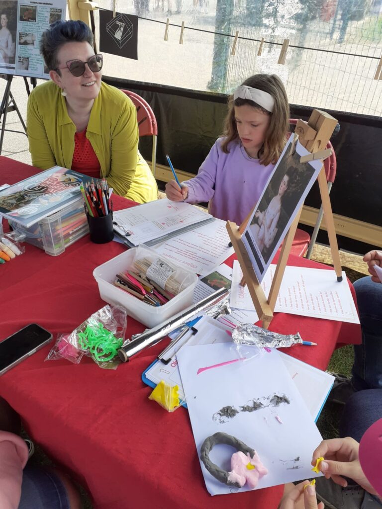 A participant sits at a red-covered activity table, with artist Laura Mabbutt, drawing on a worksheet while looking at a portrait displayed on an easel. Art materials, pencils, modelling clay and craft supplies are spread across the table, with another participant seated nearby during an Art Road Trip activity.