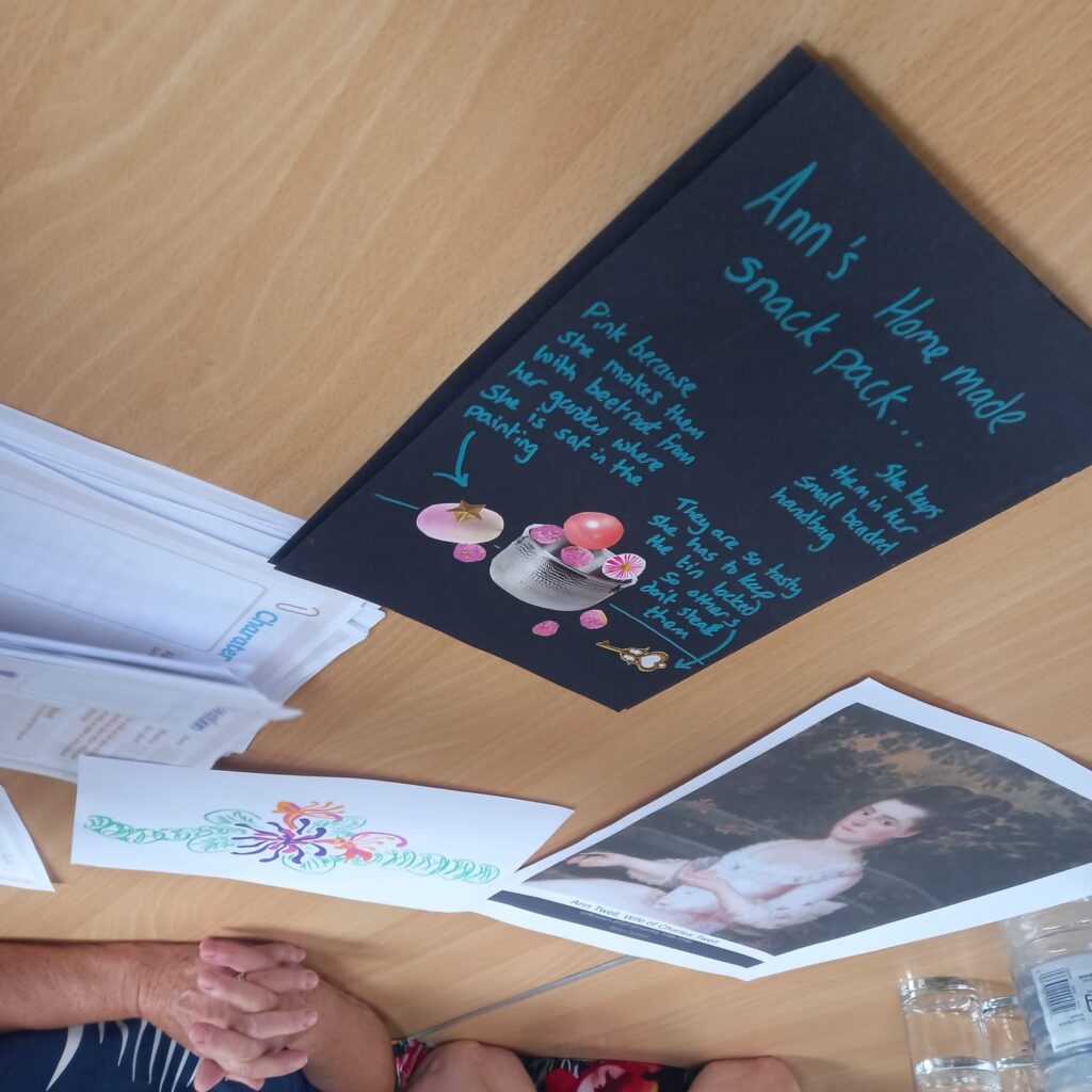 Artwork created during a workshop is displayed on a table, including a black card featuring a “snack pack” concept inspired by a portrait and a colourful floral design. Printed portrait references and activity sheets are visible alongside the artwork.