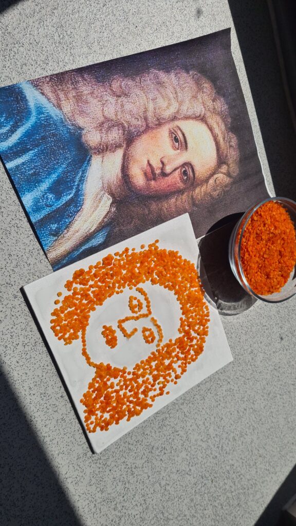 A printed portrait is paired with a seed artwork made from red lentils arranged on canvas to form the sitter’s hair and facial features. A glass container filled with lentils sits beside the finished portrait.
