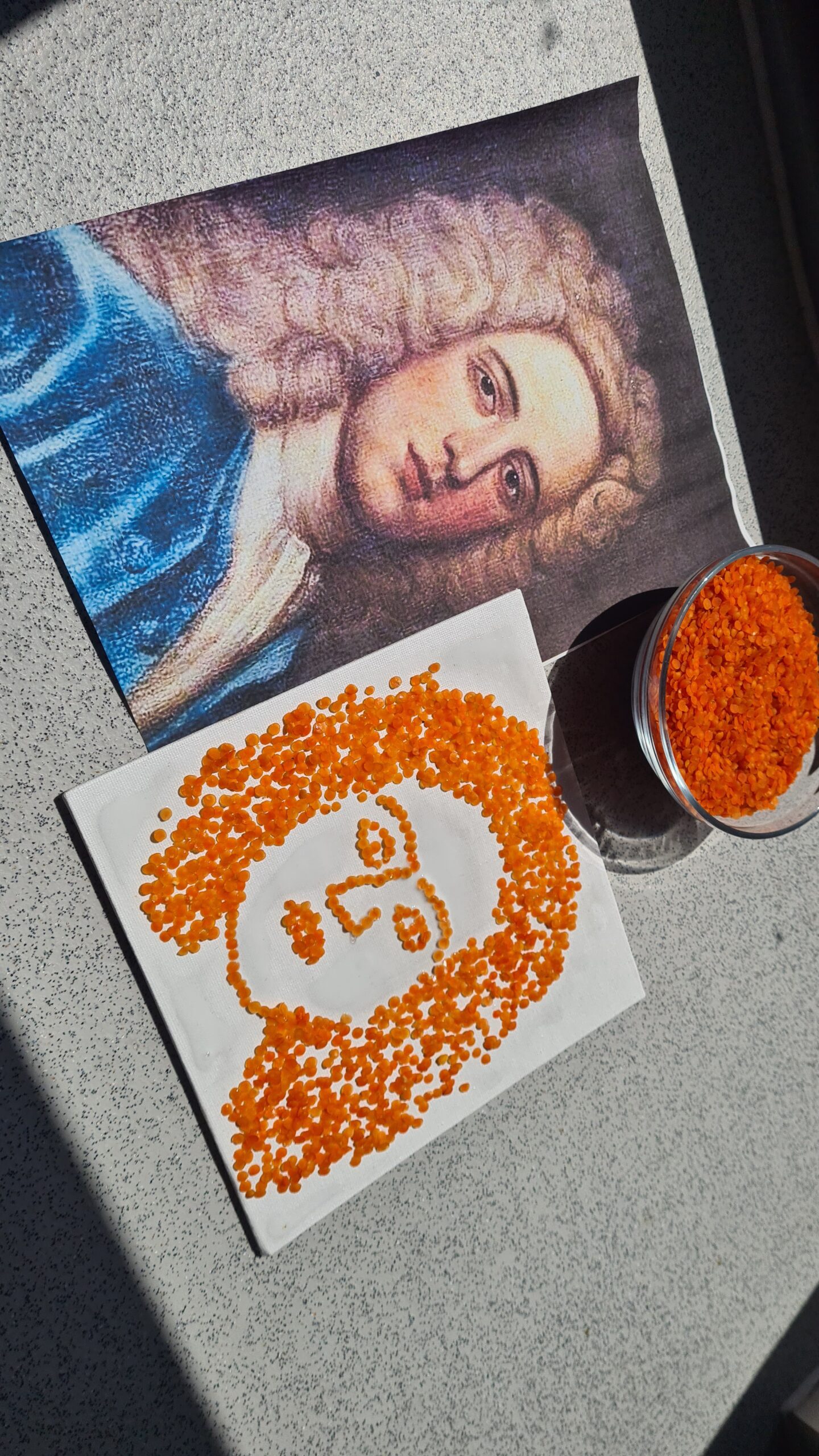 A printed portrait is paired with a seed artwork made from red lentils arranged on canvas to form the sitter’s hair and facial features. A glass container filled with lentils sits beside the finished portrait.