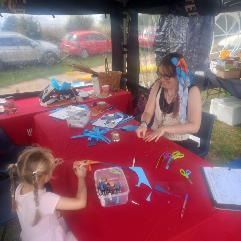 A young girl and artist Emily Cartwright sit at an outdoor table making paper headdresses inspired by Spalding's waterways