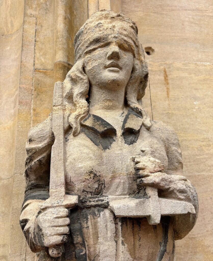 A stone, eroded statue of a blindfolded figures holds what appears to be a sword