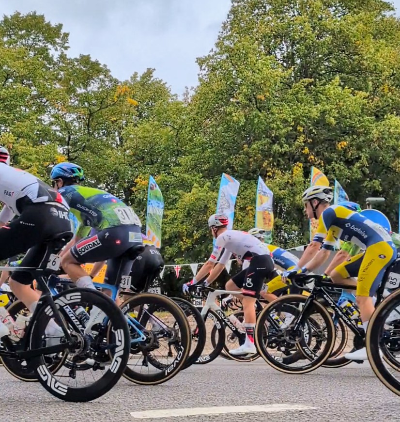 An image, taken at a low angle of Tour of Britain cyclists passing by the Transported flags