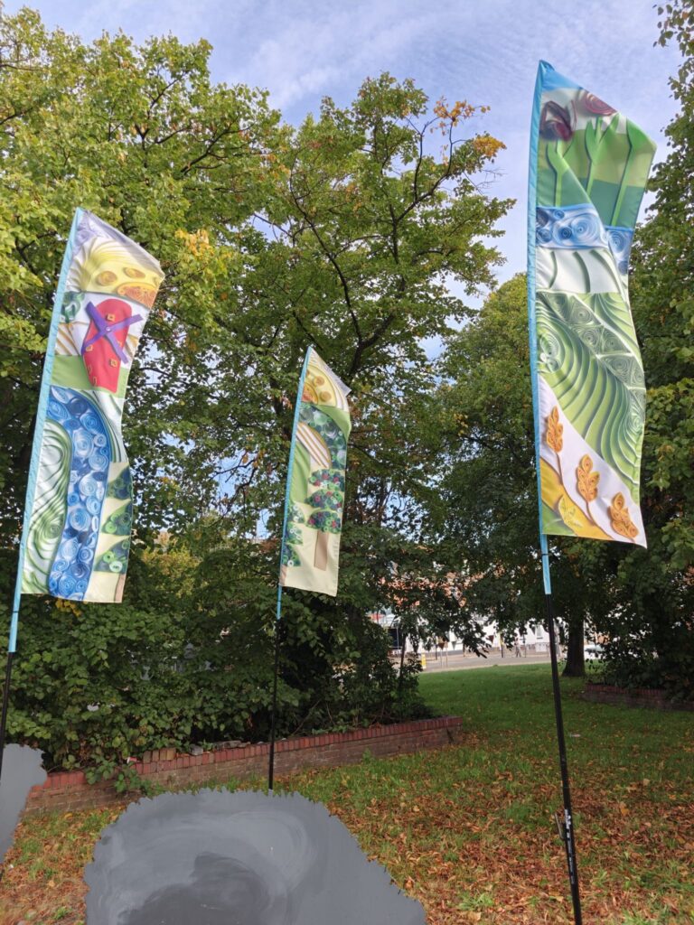 3 colourful feather flags featuring designs of fields, produce and rivers can be seen on a roadabout in Boston