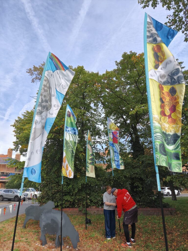 Colourful feather flags featuring designs of fields, golden wheat, produce and rivers can be seen on a roadabout in Boston with the Transported team installing them seen in the background