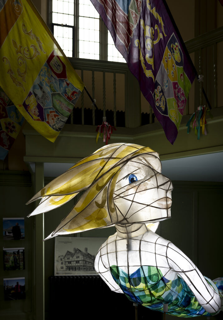 Large illuminated lantern sculpture displayed indoors beneath colourful community banners at a Transported arts event in Boston.