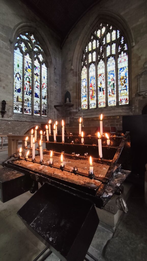 A picture of the interior of St Botolphs, showing a collection of lit prayer candles with stained glass windows beyond