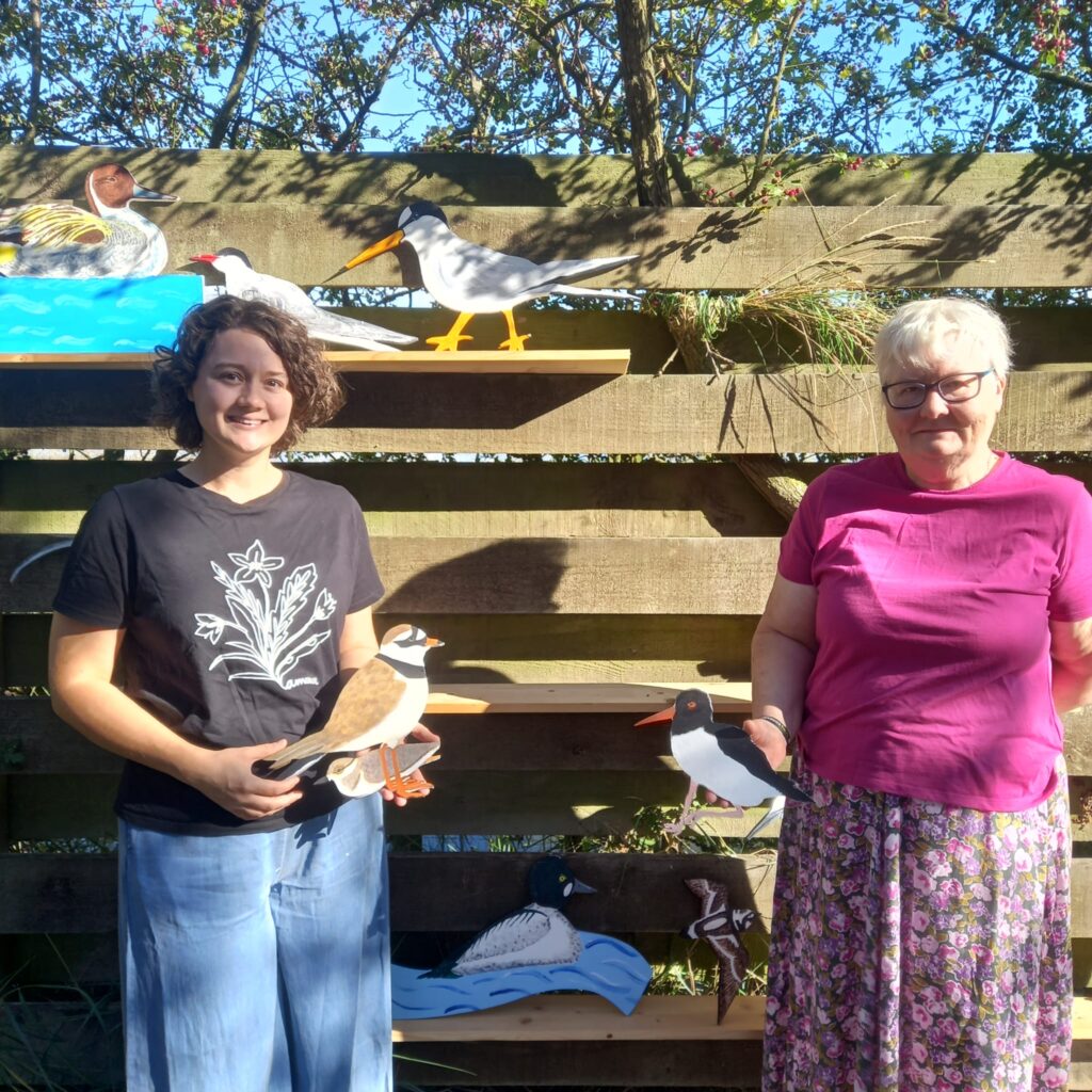 Participants smile in front of bird-shaped wooden waymarkers are fixed to a wooden pallet