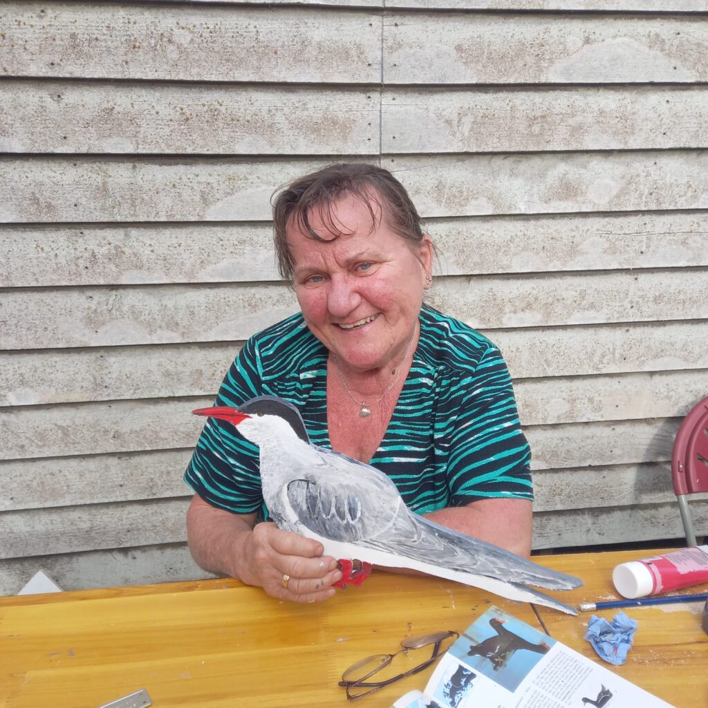 A female participant smiles and holds her hand painted bird-shaped wooden waymarker to the camera during a community art workshop at RSPB Freiston Shore.