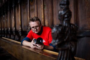 Image of photographer Steven Hatton, wearing a red Transported t-shirt, holding a camera inside a church, photographing a wooden choir stall