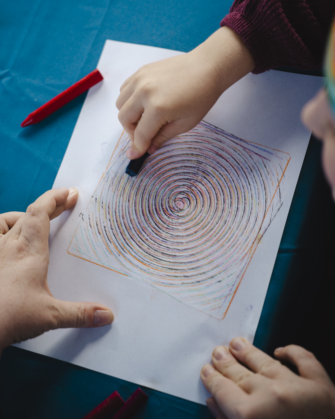 A close up image of children's hands, using crayons to create a coloured, layered spiral