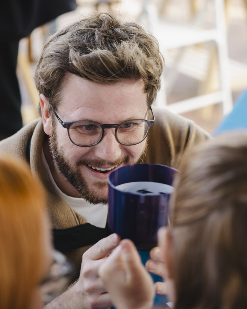 A young smiling man with dark blonde hair, glasses and a smart beard demonstrates how zoetropes work to two children