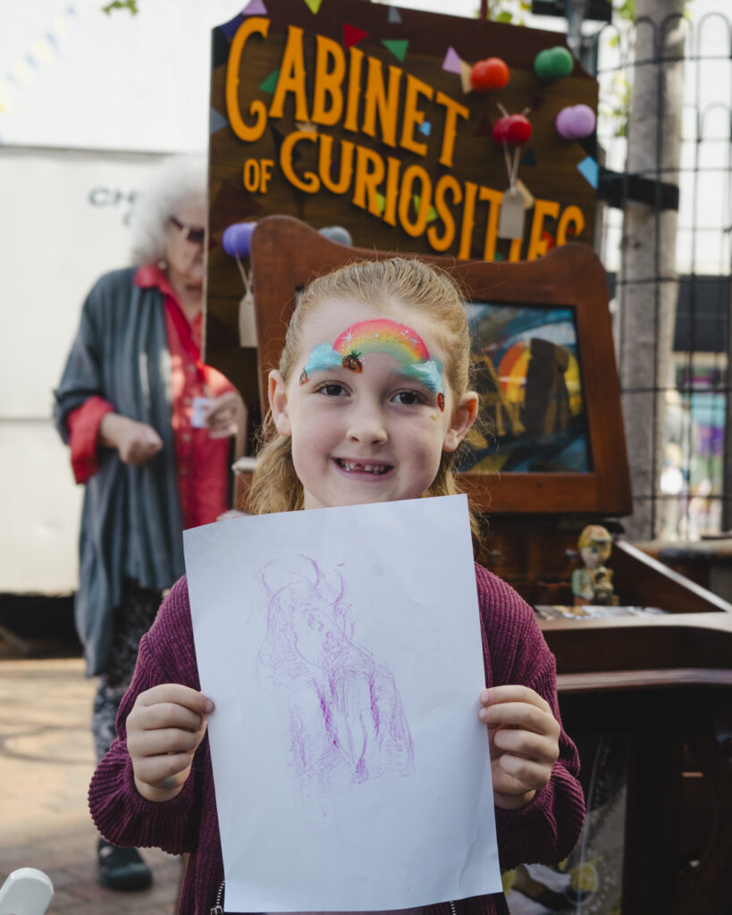 A young girl with a rainbow face painting smiles at the camera showing her brass rubbing picture in front of the Cabinet of Curiosities in Spalding