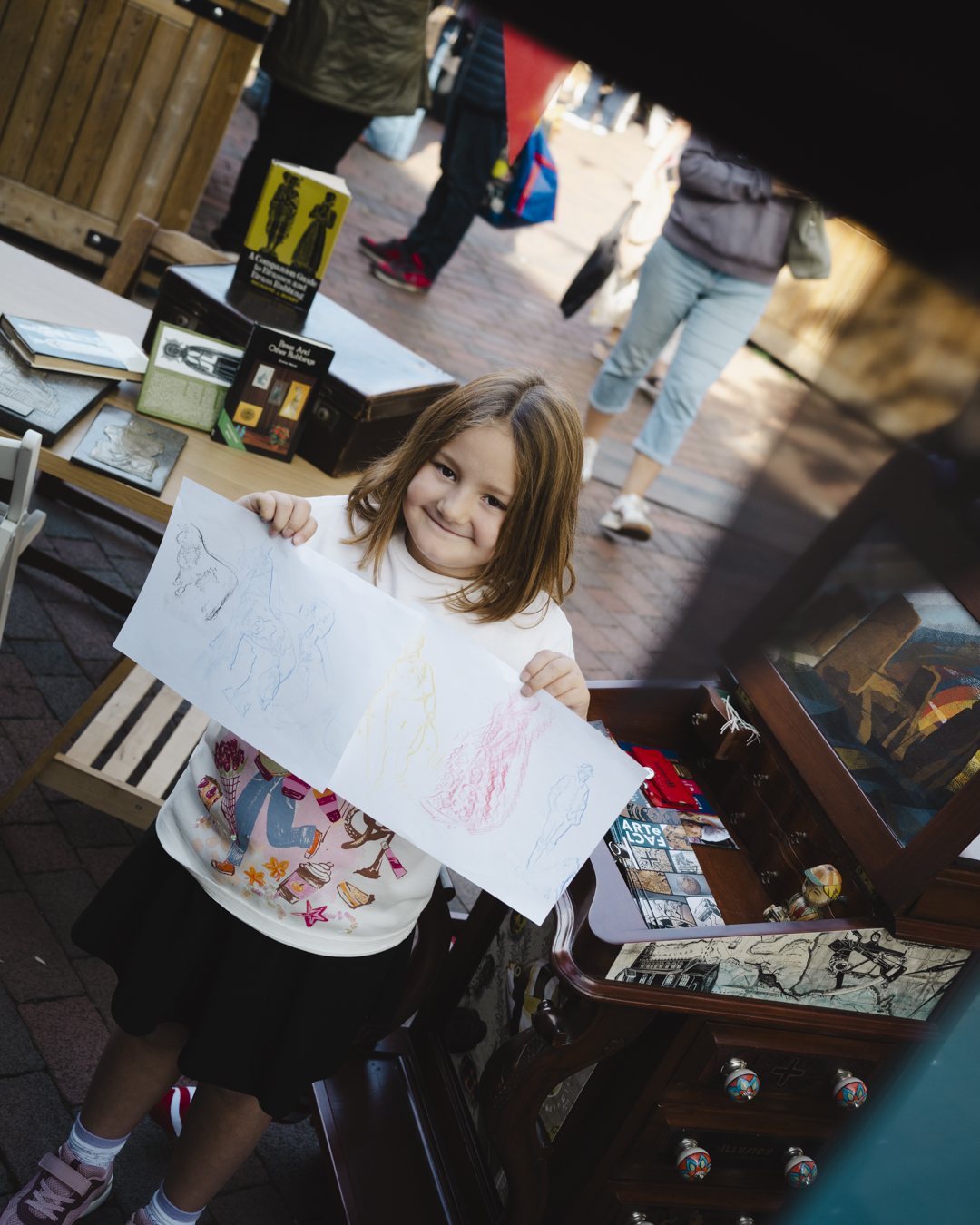 Image of a young girl stood next to the Cabinet of Curiosities holding up her brass rubbing artwork to the camera