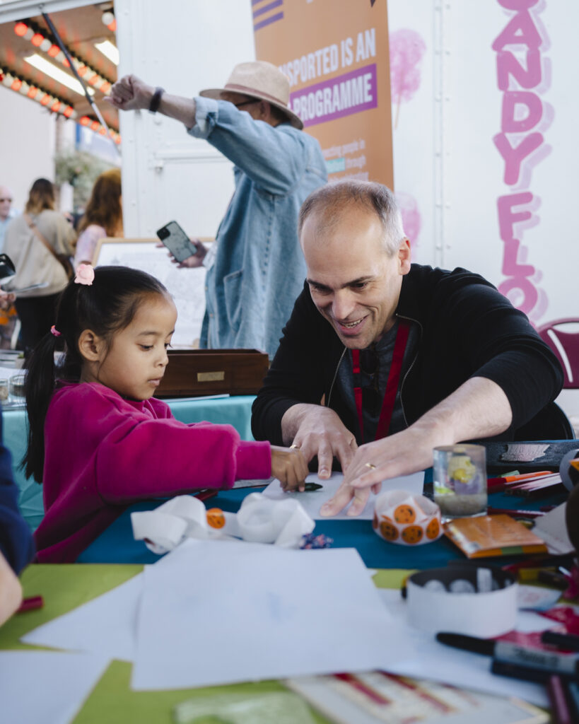 Image of a young girl in a pink jumper gets involved in an arts activity, assisted by a friendly smiling male member of the Spalding Gentleman Society's team
